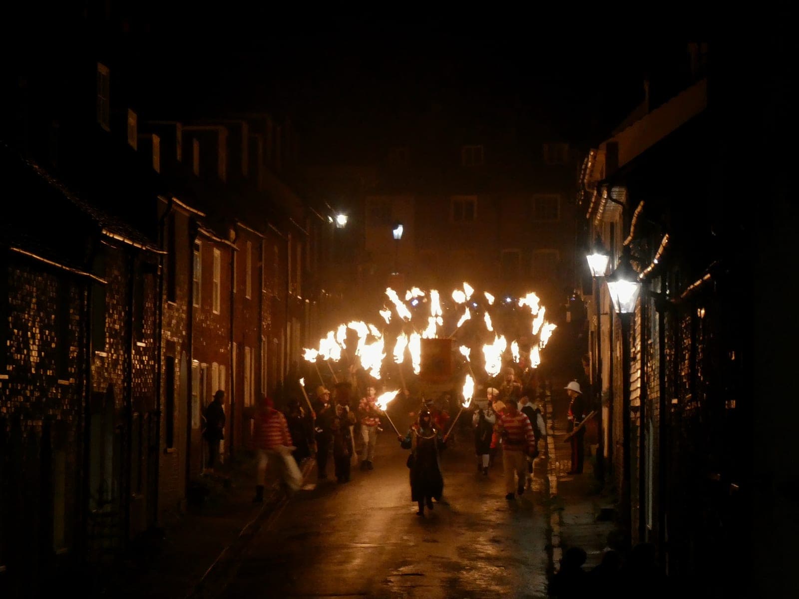 Costumed marchers in striped jumpers carry flaming torches down a narrow, rain-slicked street during the Lewes Bonfire procession at night