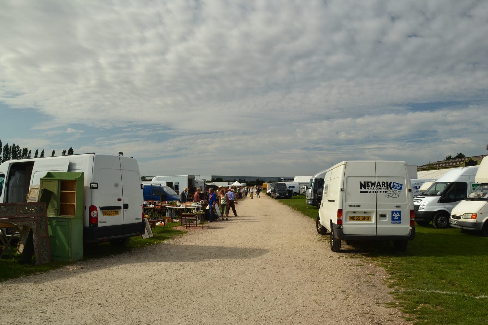 Dealers' vans backed onto grass at Newark Antiques and Collectors' Fair, with painted furniture and trestle tables of stock beside a gravel track.